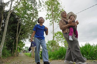 Asyalı Müslüman anne, çocuklarıyla yemyeşil bir parkta ağaçlık bir patika boyunca yürürken bebeğini taşıyor. Aile, sağlıklı bir yaşam tarzını, ebeveynliği, birlikteliği ve her şeyi temsil eden doğal bir çevrede huzurlu bir doğa yürüyüşü yapmayı seviyor.