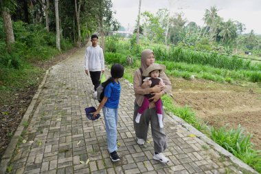 Asyalı Müslüman anne, çocuklarıyla yemyeşil bir parkta ağaçlık bir patika boyunca yürürken bebeğini taşıyor. Aile, sağlıklı bir yaşam tarzını, ebeveynliği, birlikteliği ve her şeyi temsil eden doğal bir çevrede huzurlu bir doğa yürüyüşü yapmayı seviyor.