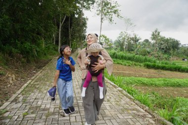 Asyalı Müslüman anne, çocuklarıyla yemyeşil bir parkta ağaçlık bir patika boyunca yürürken bebeğini taşıyor. Aile, sağlıklı bir yaşam tarzını, ebeveynliği, birlikteliği ve her şeyi temsil eden doğal bir çevrede huzurlu bir doğa yürüyüşü yapmayı seviyor.