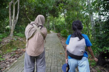 Asyalı Müslüman bir anne, çocuklarıyla birlikte yemyeşil bir parkta ağaçlık bir patika boyunca yürürken, sağlıklı yaşam tarzını, ebeveynliği, birlikteliği ve günlük aile yaşamını temsil ederek, doğal bir ortamda huzurlu bir şekilde yürümeyi seviyor.