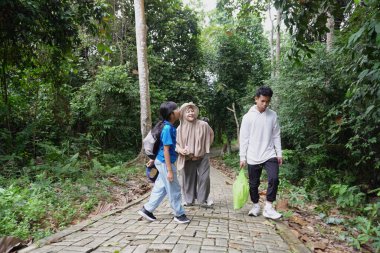Asyalı Müslüman anne, çocuklarıyla yemyeşil bir parkta ağaçlık bir patika boyunca yürürken bebeğini taşıyor. Aile, sağlıklı bir yaşam tarzını, ebeveynliği, birlikteliği ve her şeyi temsil eden doğal bir çevrede huzurlu bir doğa yürüyüşü yapmayı seviyor.