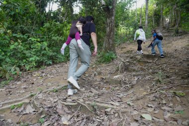 Çocuklar ilerlerken, bir çocuğun sırtında bir çocukla orman yolunda yürüyen bir aile. Sahnede açık hava maceraları, aile bağları ve verimli tropikal orman doğasıyla çevrili aktif bir yaşam tarzı var.