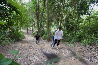 Çocuklar ilerlerken, bir çocuğun sırtında bir çocukla orman yolunda yürüyen bir aile. Sahnede açık hava maceraları, aile bağları ve verimli tropikal orman doğasıyla çevrili aktif bir yaşam tarzı var.