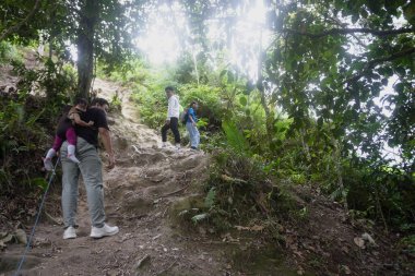 Çocuklar önlerinde yürürken, bir babayla birlikte bir orman yolunda yürüyen bir aile. Bereketli bir tropikal ormanda açık hava macerası ve aile bağları aktif bir yaşam tarzını, keşifleri ve doğadaki birlikteliği temsil eder.