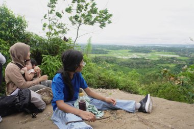 Asyalı Müslüman anne, çocuğuyla birlikte yeşil vadilere ve pirinç tarlalarına bakan manzaralı bir yamaçta dinlenirken bebeğini kucağında tutuyor. Aile, doğa ve temiz havayı bir gezide, aile seyahatini, birlikteliği ve iyileşmeyi temsil ederek sever.