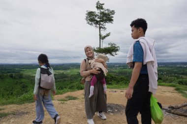Asyalı Müslüman anne, çocuğuyla birlikte yeşil vadilere ve pirinç tarlalarına bakan manzaralı bir yamaçta dinlenirken bebeğini kucağında tutuyor. Aile, doğa ve temiz havayı bir gezide, aile seyahatini, birlikteliği ve iyileşmeyi temsil ederek sever.