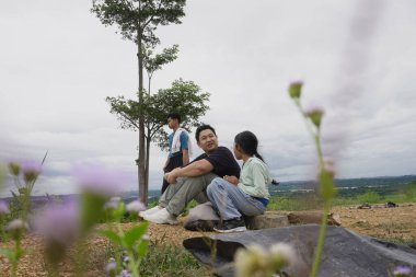 Bulutlu bir gökyüzünün altında manzaranın tadını çıkarırken bir tepede dinlenen bir aile. Bir baba kızıyla oturur, etrafı kır çiçekleri ve doğal manzaralarla çevrili, aile bağlarını, açık hava yaşam tarzını, seyahatleri ve huzurlu kırsal bölgeleri temsil eder.
