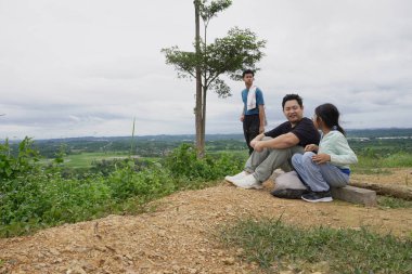 Bulutlu bir gökyüzünün altında manzaranın tadını çıkarırken bir tepede dinlenen bir aile. Bir baba kızıyla oturur, etrafı kır çiçekleri ve doğal manzaralarla çevrili, aile bağlarını, açık hava yaşam tarzını, seyahatleri ve huzurlu kırsal bölgeleri temsil eder.