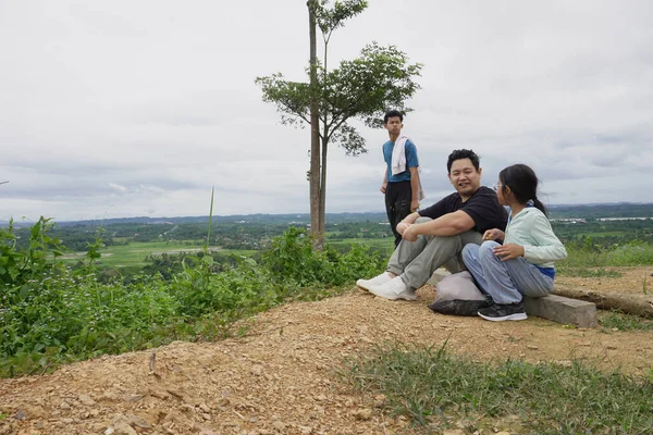 Bulutlu bir gökyüzünün altında manzaranın tadını çıkarırken bir tepede dinlenen bir aile. Bir baba kızıyla oturur, etrafı kır çiçekleri ve doğal manzaralarla çevrili, aile bağlarını, açık hava yaşam tarzını, seyahatleri ve huzurlu kırsal bölgeleri temsil eder.