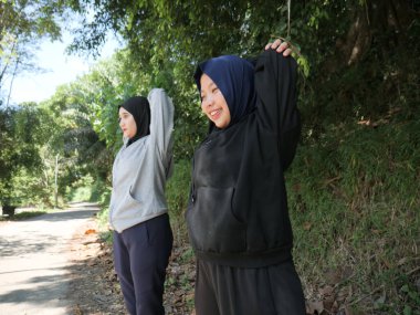 Yeşil bir parkta güneşli bir günde, tesettür takan ve ısınan iki genç Müslüman kadın. Sağlıklı yaşam tarzı, arkadaşlık ve doğal ışık ile uygunluk kavramı