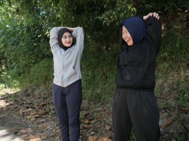 Yeşil bir parkta güneşli bir günde, tesettür takan ve ısınan iki genç Müslüman kadın. Sağlıklı yaşam tarzı, arkadaşlık ve doğal ışık ile uygunluk kavramı