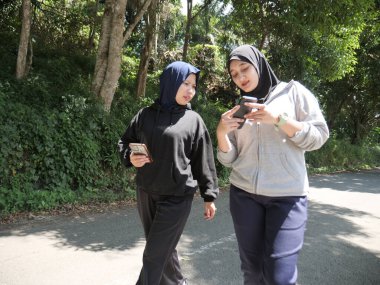 Yeşil bir parkta başörtüsü takan iki genç Müslüman kadın güneşli bir günde mutlu bir şekilde gülümseyen akıllı telefon ekranına bakıp paylaşıyorlar. Arkadaşlık, teknoloji ve doğal ışık ile modern sağlıklı yaşam tarzı kavramı.