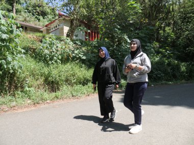 Güneşli bir günde ağaçlarla dolu yeşil bir park yolunda sabah egzersizi için birlikte yürüyen tesettür giyen iki genç Müslüman kadın. Aktif yaşam tarzı, arkadaşlık ve doğal ışık ile sağlıklı yaşam konsepti