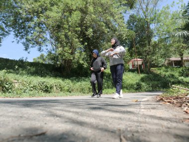 Güneşli bir günde ağaçlarla dolu yeşil bir park yolunda sabah egzersizi için birlikte yürüyen tesettür giyen iki genç Müslüman kadın. Aktif yaşam tarzı, arkadaşlık ve doğal ışık ile sağlıklı yaşam konsepti