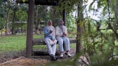 İki genç Asyalı Müslüman kız, büyük ağaçların altında, ahşap bir çardakta oturup sohbet ediyor ve sabah güneşinin altında bir orman parkında doğanın tadını çıkarıyorlar.