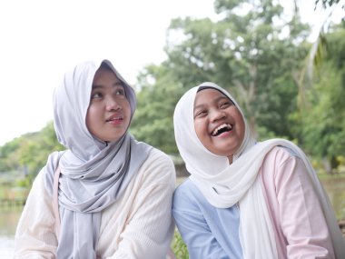 İki mutlu Asyalı Müslüman kız kardeş gün batımında gölün kenarında bir ağacın altında oturup sohbet ediyor ve doğada birlikte gülüyorlar.