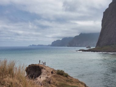 Turistli Madeira Sahil Hattı Vista - Madeira 'nın yüksek uçurumları, bulutları ve küçük turist figürleriyle dolu engebeli kıyı şeridine dramatik bir okyanus manzarası.