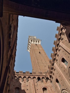 Palazzo Pubblico Avlusu 'ndan Torre del Mangia - Siena, Piazza del Campo' daki Palazzo Pubblico iç avlusunun ortaçağ mimarisi tarafından çerçevelenen yüksek kırmızı tuğlalı Torre del Mangia 'nın dramatik alçak açılı manzarası.