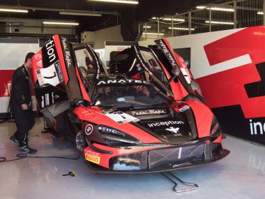 Çukur Garajındaki McLaren GT3 Yarış Arabası - Kırmızı ve siyah bir McLaren 'ın ön görüntüsü Kelebek kapıları açık, modern, aydınlık bir garajda dinleniyor.