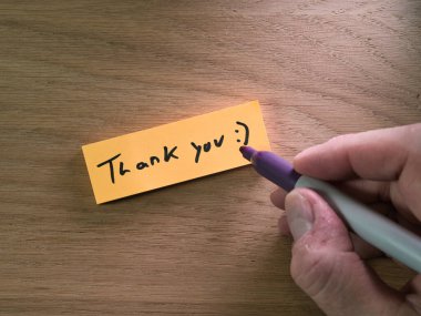 Tahta Masa 'ya Teşekkür Notu - Teşekkür ederim:)' Mor bir kalemle doğal ahşap bir masanın üzerine, minnet ve takdiri ifade eden, turuncu bir not bırakarak.