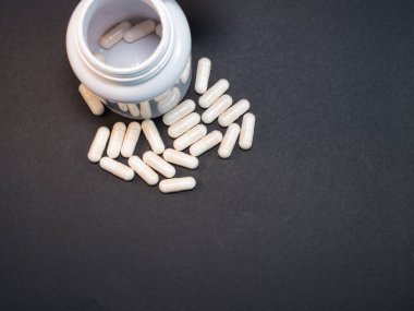 Karanlık Arkaplan 'daki ilaç kapsülleri ilaç şişesinden koyu gri arka plana beyaz ilaç kapsülleri dökülüyor. Sağlık, ilaç, tedavi ve ek maddeler kavramları. Kopyalama alanı olan düz.