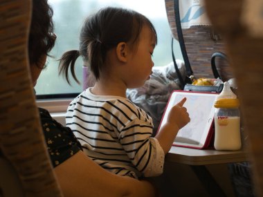 Şangay-Şangay-Çin-17 Ağustos 2018, Asyalı bir kız uzun mesafe tren yolculuğunda bir yetişkinin yanında otururken eğlenmek için dijital tablet kullanıyor. Yakınlarda bir güvercin markalı bebek şişesi duruyor..