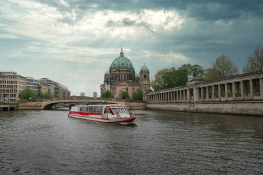 BERLIN-Almanya-18 Nisan 2025, Berlin, Almanya 'da Spree Nehri' nde gezen bir turist teknesi, ikonik Berliner Dom (Berlin Katedrali) ile dramatik bulutlu gökyüzünün altında görünür..