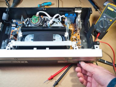 VHS bantlı VCR Onarım ve Bakım; Parçalanmış bir VCR mekanizmasını tamir eden bir teknisyenin genel görünümü. Dahili devre kartları ve test araçları ile çevrili bir VHS bandı yüklendi.