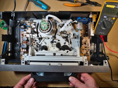 VHS bantlı VCR Onarım ve Bakım; Parçalanmış bir VCR mekanizmasını tamir eden bir teknisyenin genel görünümü. Dahili devre kartları ve test araçları ile çevrili bir VHS bandı yüklendi.