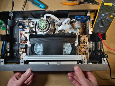 VHS bantlı VCR Onarım ve Bakım; Parçalanmış bir VCR mekanizmasını tamir eden bir teknisyenin genel görünümü. Dahili devre kartları ve test araçları ile çevrili bir VHS bandı yüklendi.