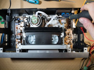 VHS bantlı VCR Onarım ve Bakım; Parçalanmış bir VCR mekanizmasını tamir eden bir teknisyenin genel görünümü. Dahili devre kartları ve test araçları ile çevrili bir VHS bandı yüklendi.
