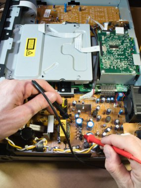 Eller Elektronik Devre Tahtasını Çokölçer ile Test Ediyor; Açık bir DVD / Blu-ray oynatıcı veya benzeri bir aygıt içindeki kompleks elektronik devreleri tanımlamak veya onarmak için çokölçer kullanan teknisyenlerin ellerinin yakın çekimi.