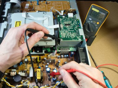 Eller Elektronik Devre Tahtasını Çokölçer ile Test Ediyor; Açık bir DVD / Blu-ray oynatıcı veya benzeri bir aygıt içindeki kompleks elektronik devreleri tanımlamak veya onarmak için çokölçer kullanan teknisyenlerin ellerinin yakın çekimi.