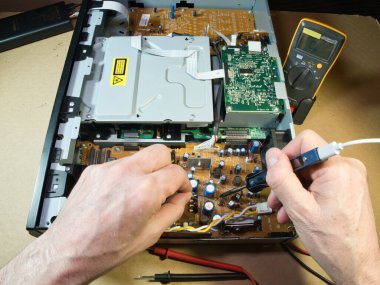 Eller Elektronik Devre Tahtasını Çokölçer ile Test Ediyor; Açık bir DVD / Blu-ray oynatıcı veya benzeri bir aygıt içindeki kompleks elektronik devreleri tanımlamak veya onarmak için çokölçer kullanan teknisyenlerin ellerinin yakın çekimi.