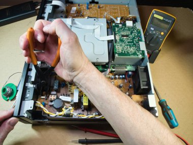 Eller Elektronik Devre Tahtasını Çokölçer ile Test Ediyor; Açık bir DVD / Blu-ray oynatıcı veya benzeri bir aygıt içindeki kompleks elektronik devreleri tanımlamak veya onarmak için çokölçer kullanan teknisyenlerin ellerinin yakın çekimi.