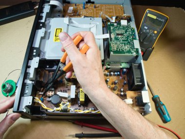 Eller Elektronik Devre Tahtasını Çokölçer ile Test Ediyor; Açık bir DVD / Blu-ray oynatıcı veya benzeri bir aygıt içindeki kompleks elektronik devreleri tanımlamak veya onarmak için çokölçer kullanan teknisyenlerin ellerinin yakın çekimi.