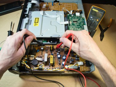 Eller Elektronik Devre Tahtasını Çokölçer ile Test Ediyor; Açık bir DVD / Blu-ray oynatıcı veya benzeri bir aygıt içindeki kompleks elektronik devreleri tanımlamak veya onarmak için çokölçer kullanan teknisyenlerin ellerinin yakın çekimi.