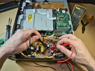 Eller Elektronik Devre Tahtasını Çokölçer ile Test Ediyor; Açık bir DVD / Blu-ray oynatıcı veya benzeri bir aygıt içindeki kompleks elektronik devreleri tanımlamak veya onarmak için çokölçer kullanan teknisyenlerin ellerinin yakın çekimi.