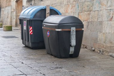 Eski Duvara Karşı Modern Sokak Atık Konteynırları; İki büyük, siyah ve mavi plastik çöp konteynırları Islak, kaldırım taşı bir cadde üzerinde Avrupa 'da antik bir taş duvarın yanında.