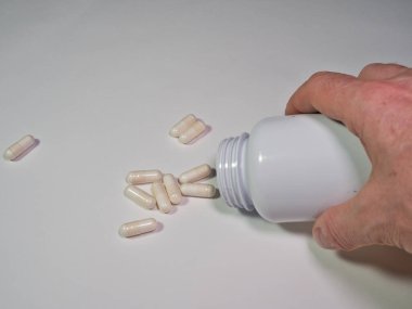 Bir el beyaz ilaç şişesinden el kapsülleri dökme; bir el beyaz bir hap şişesini devirir, açık renkli tıbbi kapsülleri nötr bir arka plan üzerine döker. Sağlık kavramları, aşırı doz ve günlük takviyeler.