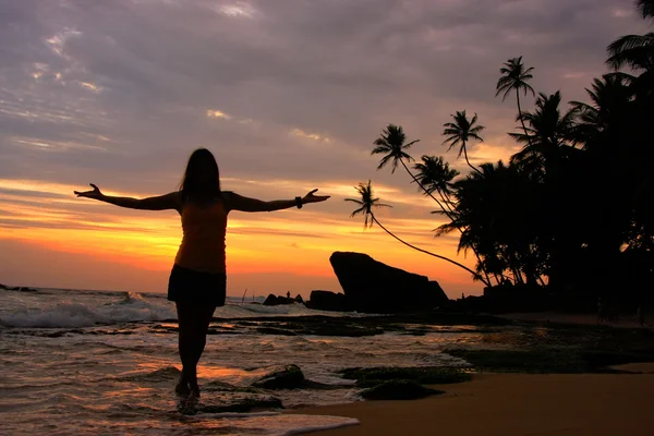 Silhouetted woman on a beach with palm trees and rocks at sunset — Stock Photo, Image