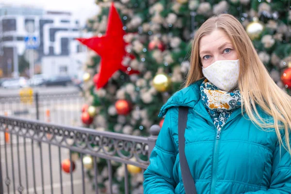 Bir köknar ağacının arka planında koruyucu maskeli sarışın bir kadın. Koronavirüs salgını sırasında Noel ve yeni yılı kutluyoruz.