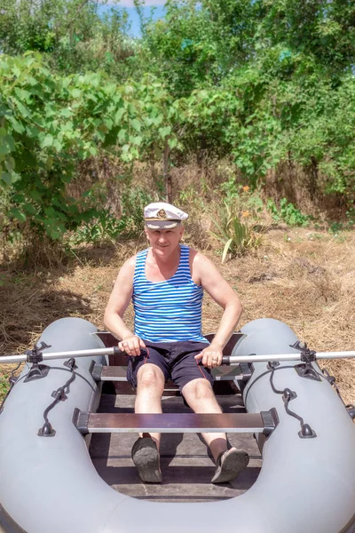 A man in a naval cap sits in a rubber boat standing on the grass. A retired sailor misses the sea. Maintenance and operation of rubber inflatable swimming facilities — Stock Image Man Naval Cap Sits Rubber Boat Standing Grass Retired Sailor Royalty Free Stock Photos