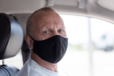 European adult man in a black protective mask is sitting in a car interior. The epidemic and lifestyle of people. clipart