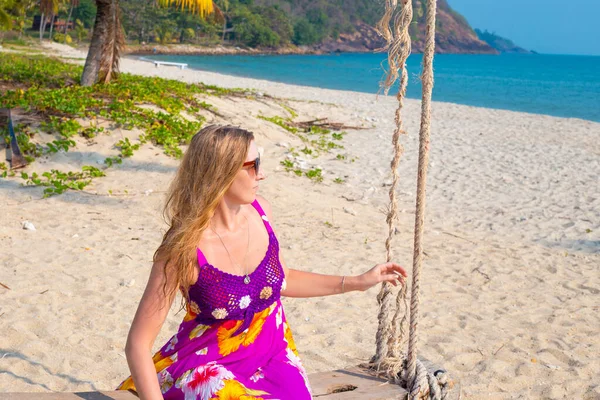 woman in a bright dress with a floral print sits on a swing and looks at the sea, tropical holidays on the islands in Asia, beautiful nature