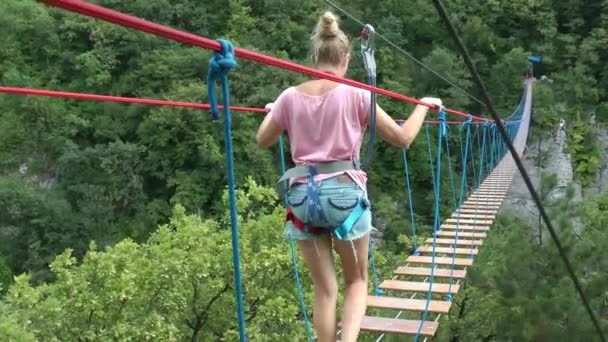 Uma menina em equipamentos de segurança caminha ao longo de uma ponte suspensa corda sobre — Vídeo de Stock