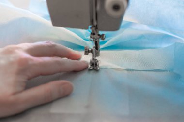 Tıbbi koruyucu giysileri ayak dikiş makinesiyle dikmek, tıbbi mavi kumaştan ayakkabı örtüleri dikmek, koronavirüsten korunma eksikliği.