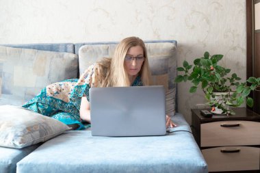 Avrupalı uzun saçlı, gözlüklü sarışın bir kadın kanepeye uzanıyor ve dizüstü bilgisayarın ekranına bakıyor, evde kendini karantinaya almış, uzaktan kumandalı.