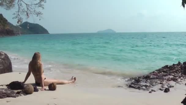 Una rubia de pelo largo en bikini se sienta en la playa en la arena y mira al mar — Vídeo de stock