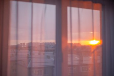 Sabah güneşinin ışınları penceredeki beyaz tüllerden düşer. Arka planda şehrin üzerinde gün doğumu. Yeni gün, atmosfer havası..